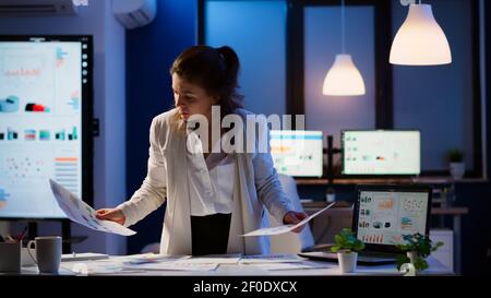 Stressed manager woman working with financial documents checking graphs, holding papers, reading raports, standing at desk late at night in start-up office doing overtime to respect deadline. Stock Photo