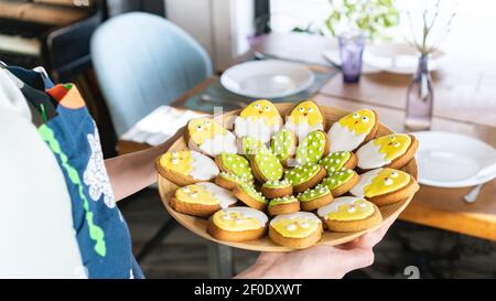 Festive table setting. A woman in an apron carries a platter of glazed Easter cookies in the shape of cute chicken-in-shell. Family dinner with sweets Stock Photo