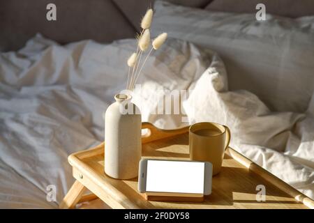 Cozy breakfast in bed. Cup of coffee, smartphone mockup, dried flowers in vase on portable wooden desk. White striped blanket and pillow on background Stock Photo