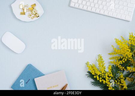 Women's desktop close-up of keyboard, notebooks, mouse and yellow branches of mimosa flowers. Flat lay.  Background, copy space. Stock Photo