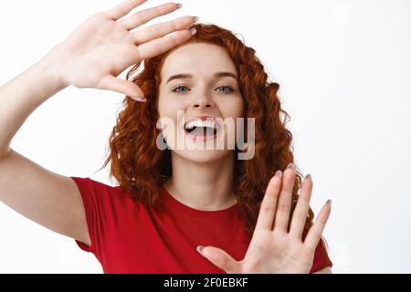 Close-up portrait of happy redhead woman making hands frame camera gesture, look through to picture or imaging something, standing in t-shirt against Stock Photo
