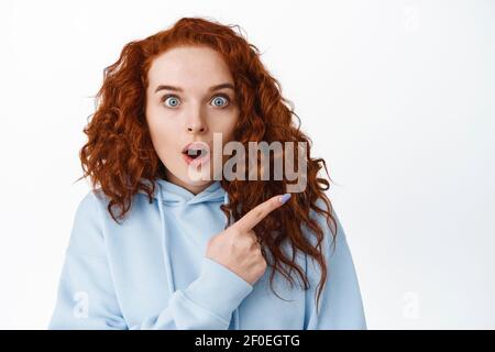 Close up of impressed teen redhead girl gasping amazed, pointing finger right at logo banner and stare at camera astonished, white background Stock Photo