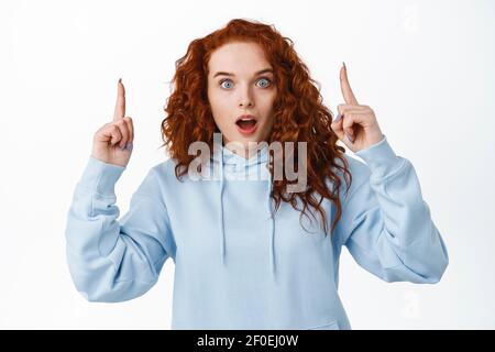 Impressed redhead girl with curly hairstyle pointing fingers up at top advertisement, gasping amazed and stare at camera with excited face, white Stock Photo