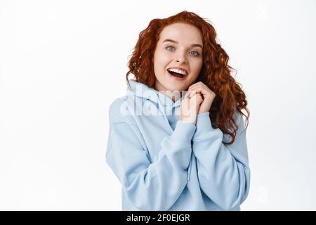 Heartfelt redhead girl look passionate and excited, holding hands near face and smiling amazed, gazing at something beautiful and touching, white Stock Photo