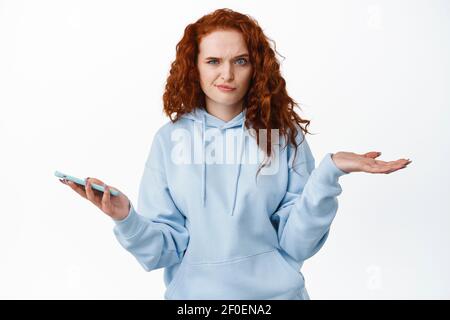 Portrait of confused and puzzled redhead girl in hoodie, holding smartphone and shrugging shoulders with frowned questioned face expression, white Stock Photo
