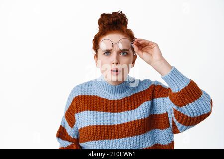 Portrait of confused redhead girl takes-off her glasses and squinting at camera, seeing something strange in distance, express surprise, white Stock Photo