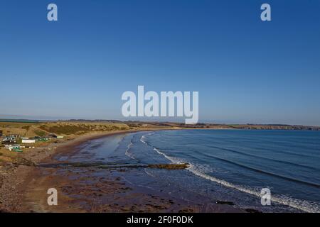 The small community of Korbie Knowe sheltered below the Cliffs at the southern end of lunan Bay facing the sandy beach with the tide going out. Stock Photo