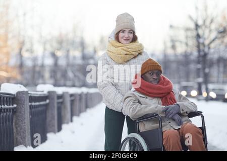 Portrait of African American man using wheelchair looking at camera while posing outdoors in winter with smiling young woman assisting, copy space Stock Photo