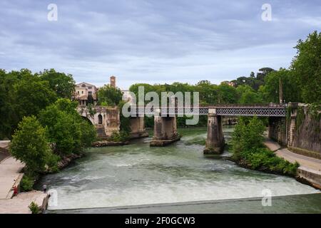The water of the Tiber river flows under the Palatine bridge, lapping the old Ponte Rotto (Emilio) bridge, green trees surround the banks of the river