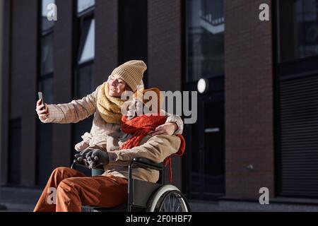 Portrait of African American man using wheelchair and taking selfie photo with young woman assisting outdoors in city, copy space Stock Photo