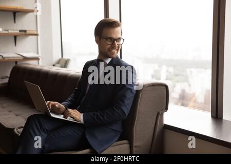 Smiling businessman wearing glasses distracted from laptop, sitting on couch Stock Photo