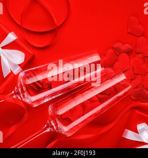 Two glass elegant champagne glasses on a bright red background with satin hearts and gifts with white bows. Close-up, top view. Stock Photo