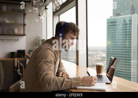 Side view motivated man wearing headphones taking notes, using laptop Stock Photo