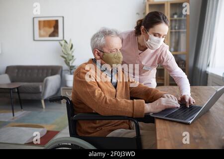 Portrait of senior man in wheelchair using laptop with nurse helping him, both wearing masks, copy space Stock Photo