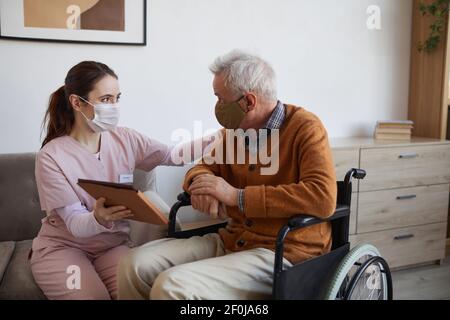 Portrait of young nurse assisting senior man in wheelchair using digital tablet at retirement home, both wearing masks, copy space Stock Photo