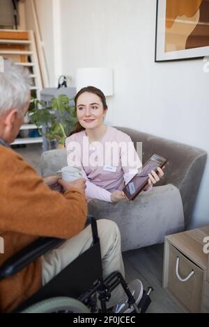 Vertical portrait of smiling female nurse talking to senior man in wheelchair while using digital tablet at retirement home Stock Photo