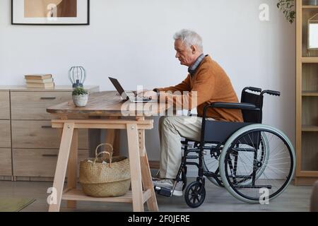 Full length side view portrait of senior man in wheelchair using laptop at home, copy space Stock Photo