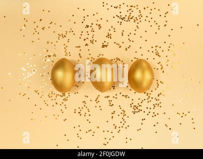 Three easter wooden golden eggs and confetti on beige backdrop. Stock Photo