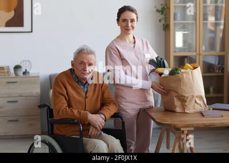 Portrait of senior man in wheelchair posing with young female caregiver holding bag of groceries, both looking at camera, copy space Stock Photo