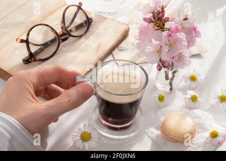 Black coffee in a cup held in the hand, next to a book with spring flowers on the table Stock Photo