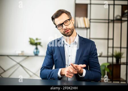 Young stately male freelancer sitting at desk in office, interviews, talking at camera, leader of the team, explaining his position or idea, conducts business training, sharing his experience Stock Photo