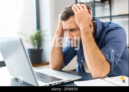 Terrified young adult businessman terrified looking at computer screen, frowning, holding the head with her hands, squints eyes, computer crashed, documents did't save, worried about financial crisis Stock Photo