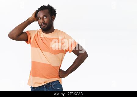 Akward and doubtful young questioned bearded african-american man in striped t-shirt standing clueless and hesitant, look away nervously, scarching Stock Photo
