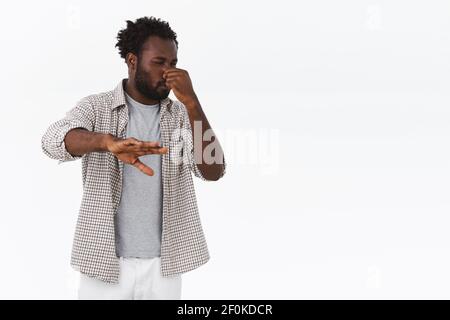 Guy disgusted from bad smell, step back and shield himself with hand. African-american bearded guy cover nose and squinting, griamace from stink or Stock Photo
