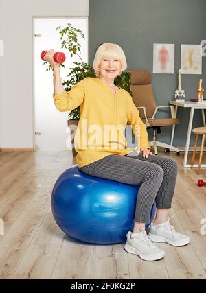 Senior woman with a dumbbell in hand sitting on a fitness ball during exercise therapy in a rehabilitation center Stock Photo
