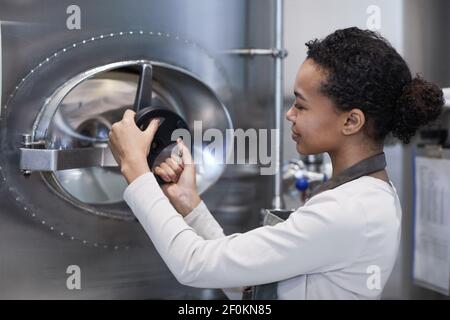 Side view portrait of smiling female worker opening hatch at storage tank in industrial workshop, copy space Stock Photo