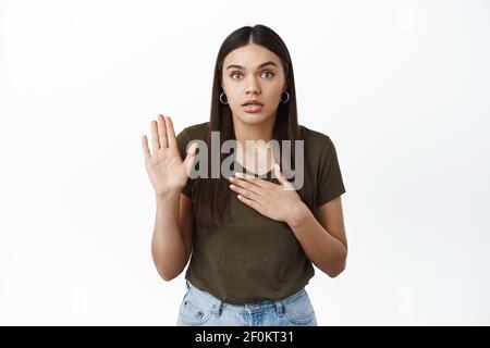 I swear its true. Young brunette woman giving oath, raising one hand and holding palm on heart, making promise, pledge to someone, standing against Stock Photo