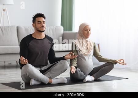 Pregnant muslim couple practicing prenatal yoga at home, meditating together Stock Photo