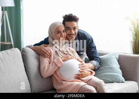 Pregnant muslim couple embracing at home, sitting on couch in living room Stock Photo