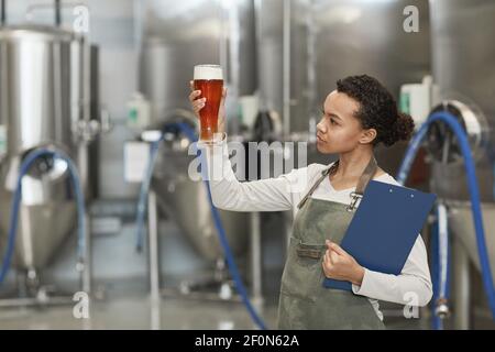 Waist up portrait of young African-American woman holding beer glass while inspecting quality of production at brewing factory, copy space Stock Photo