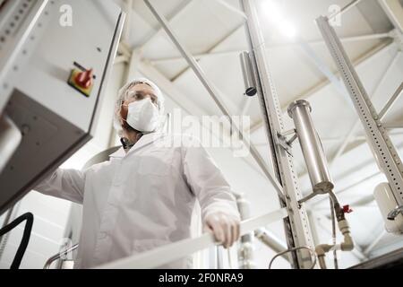 Low angle portrait of male worker operating machines at chemical plant while wearing lab coat and protective clothing, copy space Stock Photo