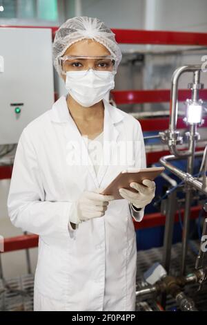 Vertical portrait of young female worker wearing protective clothing and looking at camera while using tablet at chemical plant Stock Photo