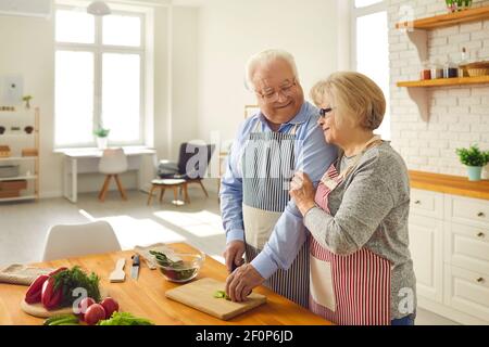Happy senior husband and wife helping each other while cooking healthy lunch at home Stock Photo