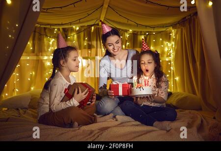 Little girl blows out candles on a cake while sitting with mom and sister in a cozy tent bed at home Stock Photo
