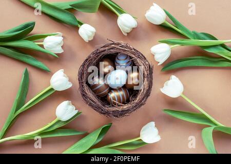 Festive Easter background of nest with painted eggs and white tulips on beige table, top view, flat lay. Spring concept. Stock Photo