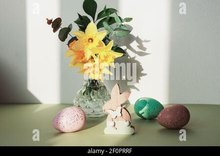 Wooden toy bunny and painted eggs, spring flowers on a green table against white wall with shadows. Easter concept. Front view. Stock Photo