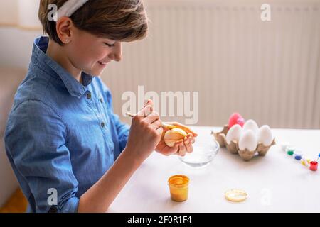 Beautiful nine-year-old girl in blue shirt paints eggs at a white table. Easter concept. Close up. Stock Photo