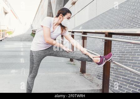 Woman with face mask dressed in sportswear stretching one leg on a railing of some stairs in the street Stock Photo