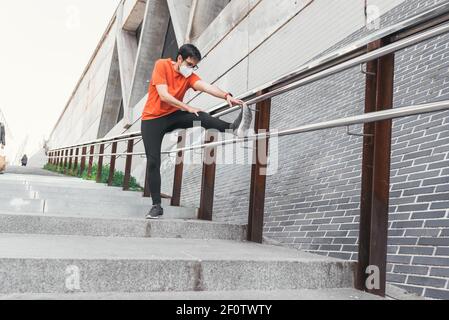 Man dressed in sportswear with face mask stretching one leg on a railing of some stairs in the street Stock Photo