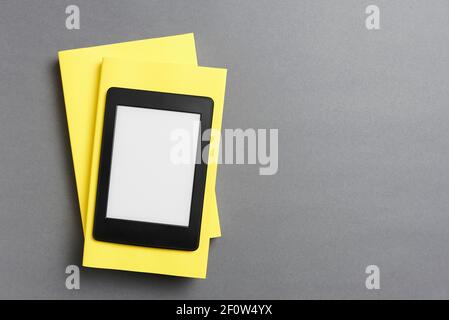 Electronic reader with with blank screen over a pair of closed yellow books, on a gray background. Concepts of technology, modernity and reading. Flat Stock Photo