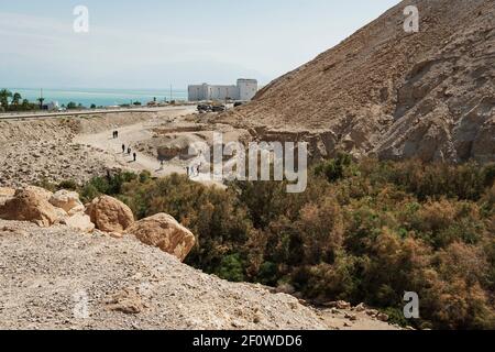 a group of hikers and tourists entering the Nahal Boqeq stream bed nature reserve in Israel with a resort hotel and the Dead Sea in the background Stock Photo
