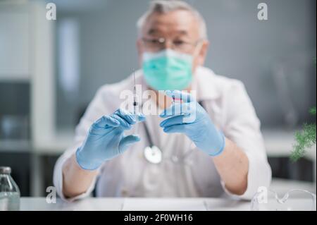 An elderly, gray-haired doctor in a spacious, bright office holds a vaccine against the covid - 19 virus strain and a syringe. Demonstrates a new drug Stock Photo