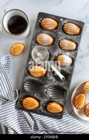 Flat lay with madeleine cakes in a baking tin, coffee a plate of cakes and a tea towel on white marble surface Stock Photo