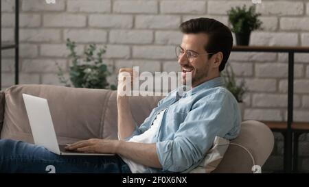 Excited young guy use laptop on sofa show winner sign Stock Photo