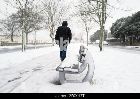 A stone and wood bench covered in snow in the central section of a tree lined street as a man walks by, it is snowing. Stock Photo