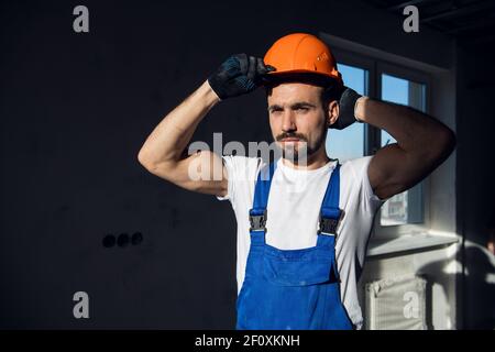 Serious man in work clothes straightens orange hard hat Stock Photo